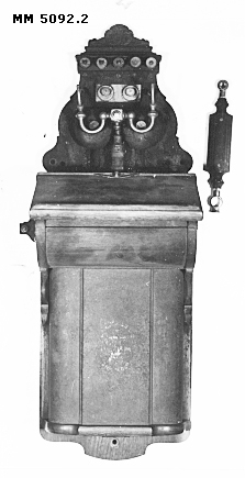 Telefon, M/1895, väggapparat, större. L.M. Ericssons tillverkning.
Apparaten är innesluten i en kåpa av mahogny. Klyka för handmikrotelefonen är placerad överst på apparaten.