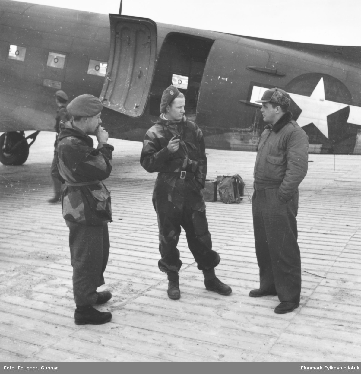 På Banak flyplass. Tre menn står og prater, to er soldater i uniform. Bak mennene står et militærfly, en C-47, Dakota.