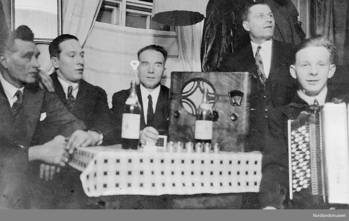 Gruppebilde av 5 menn. 4 menn sitter ved et bord, mens 1 mann står ved siden av. Mannen til høyre holder et trekkspill. Mennene er kledd i mørke jakker, lyse skjorter og mørke slips.