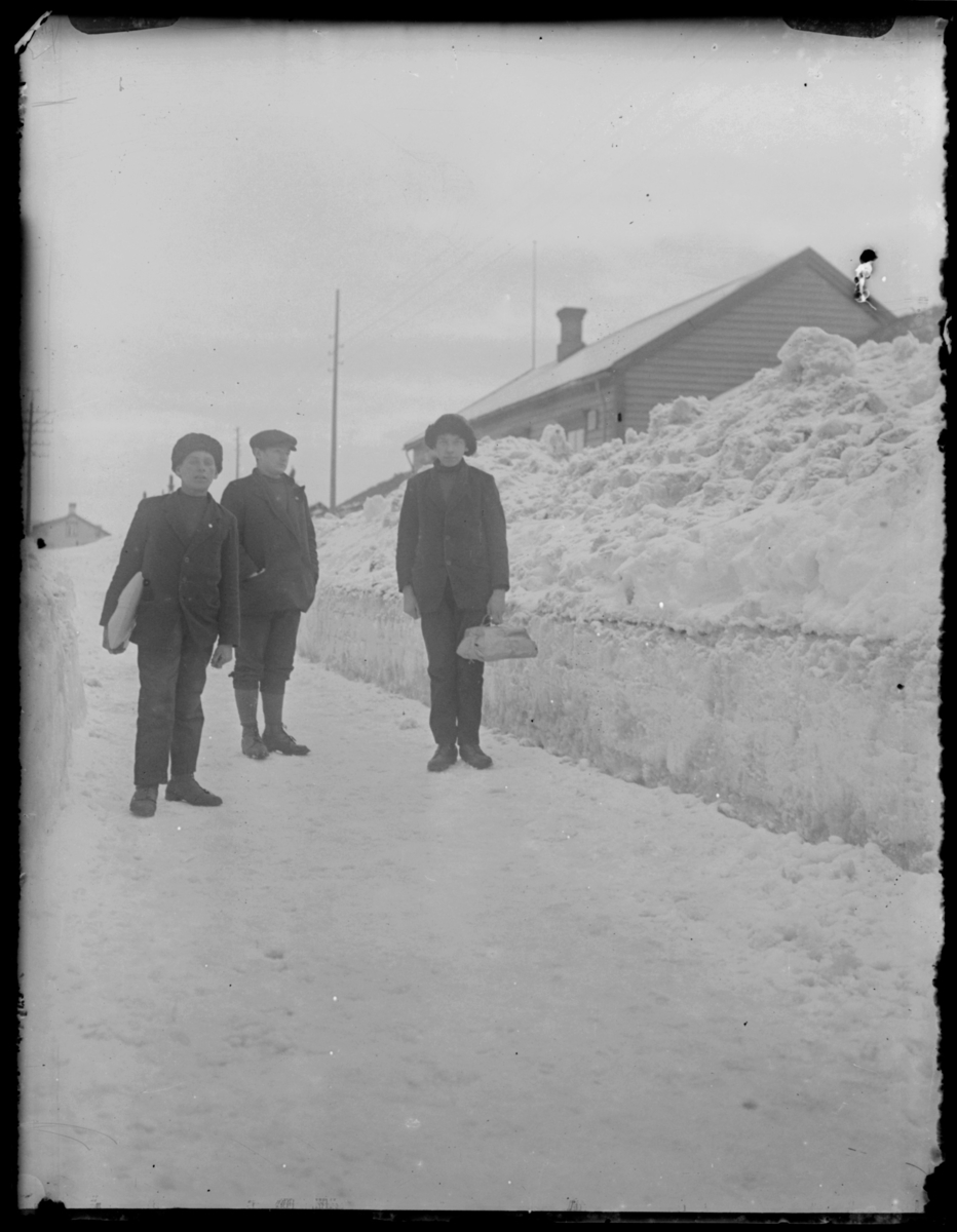Tre unge menn står på en vei, to av dem bærer pakker. Det er høye brøytekanter. To er kledt i langbukser og skinnluer mens den tredje har knebukser og caps