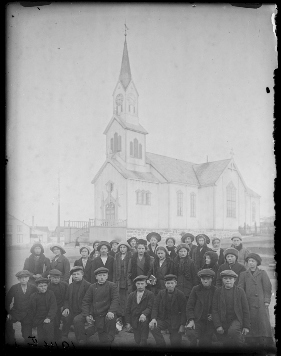 Konfirmanter i Vardø 1914, fotografert med Vardø kirke i bakgrunnen. Guttene sitter på kne foran, jentene står bak. Samme gruppe som FB 92144-002 til 007