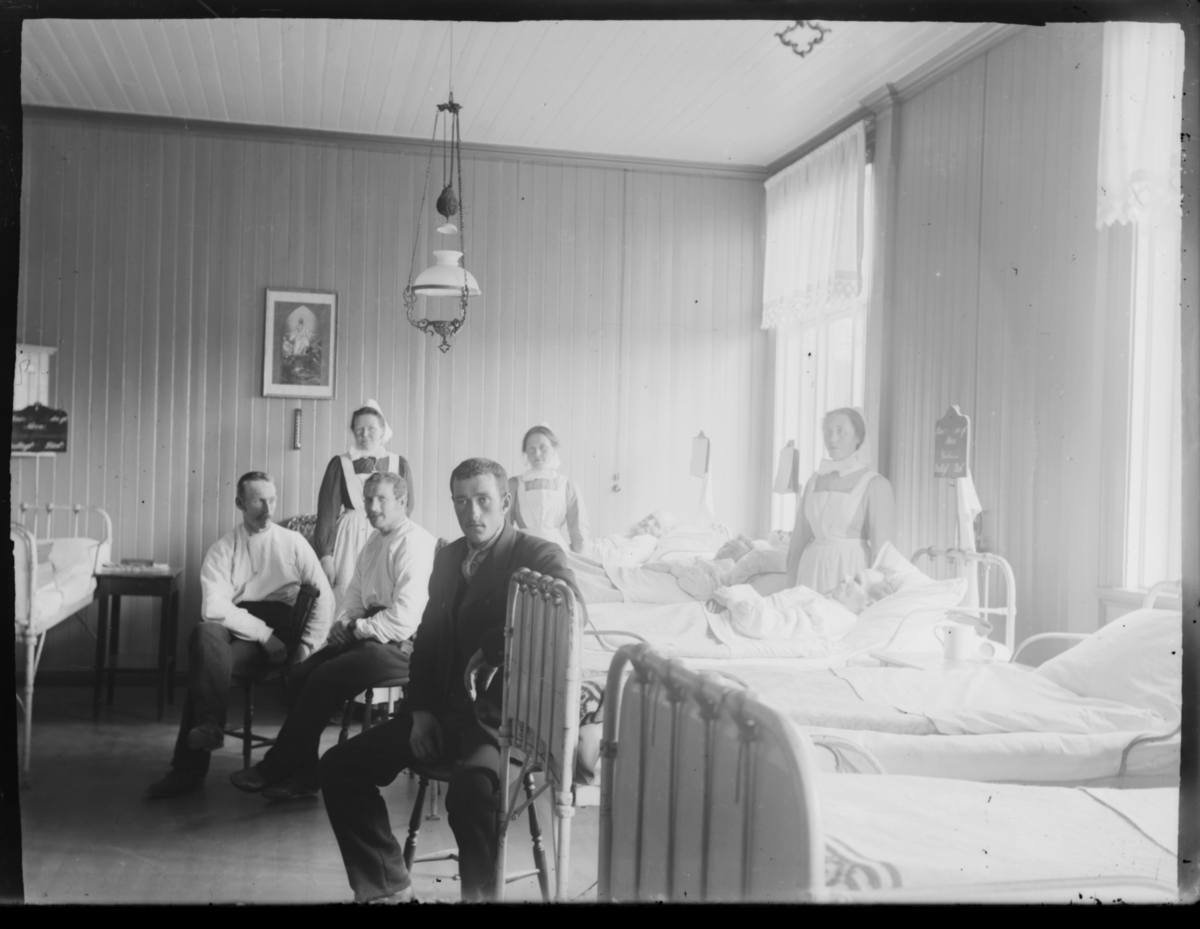 Inne på sykehuset i Vardø. Tre pasienter ligger i sengene. Tre menn sitter på krakker. Tre diakonisser (sykepleiere) står ved sengene. Det henger en lampe i taket og religiøse bilder på veggene. Også et termometer på veggen