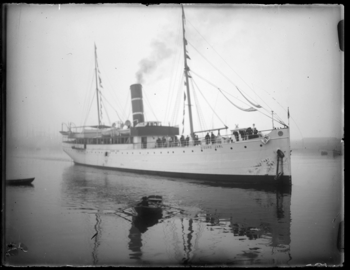 Hurtigruta 'Sigurd Jarl' ligger i havn, trolig i Vardø. Passasjerer står ved rekka. En robåt ligger ved siden av båten. Det er tåke