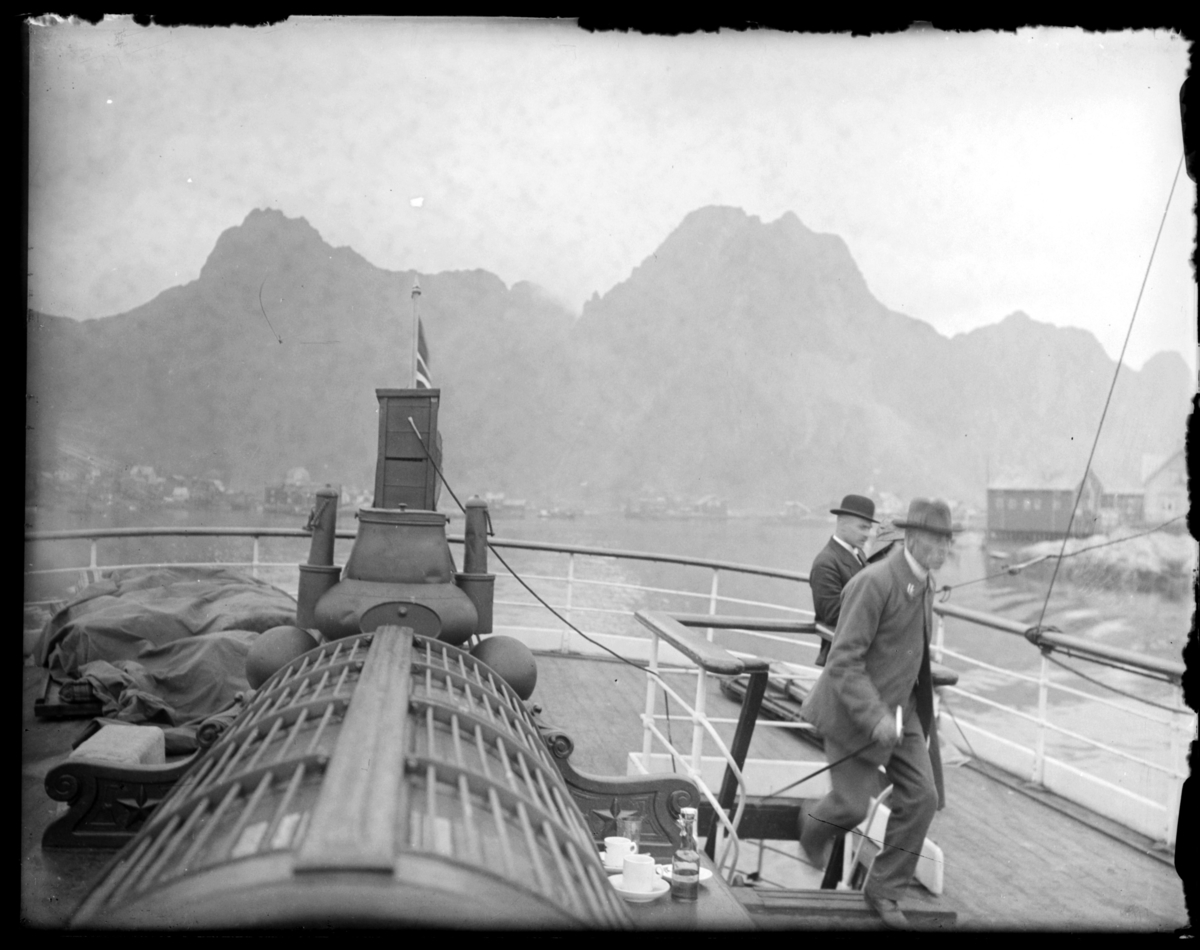 Bildet er trolig tatt fra Hurtigruta, som legger ut fra Svolvær. På en benk på dekk står to kaffekopper og en flaske. To menn på dekk. Se også FB 93149-004