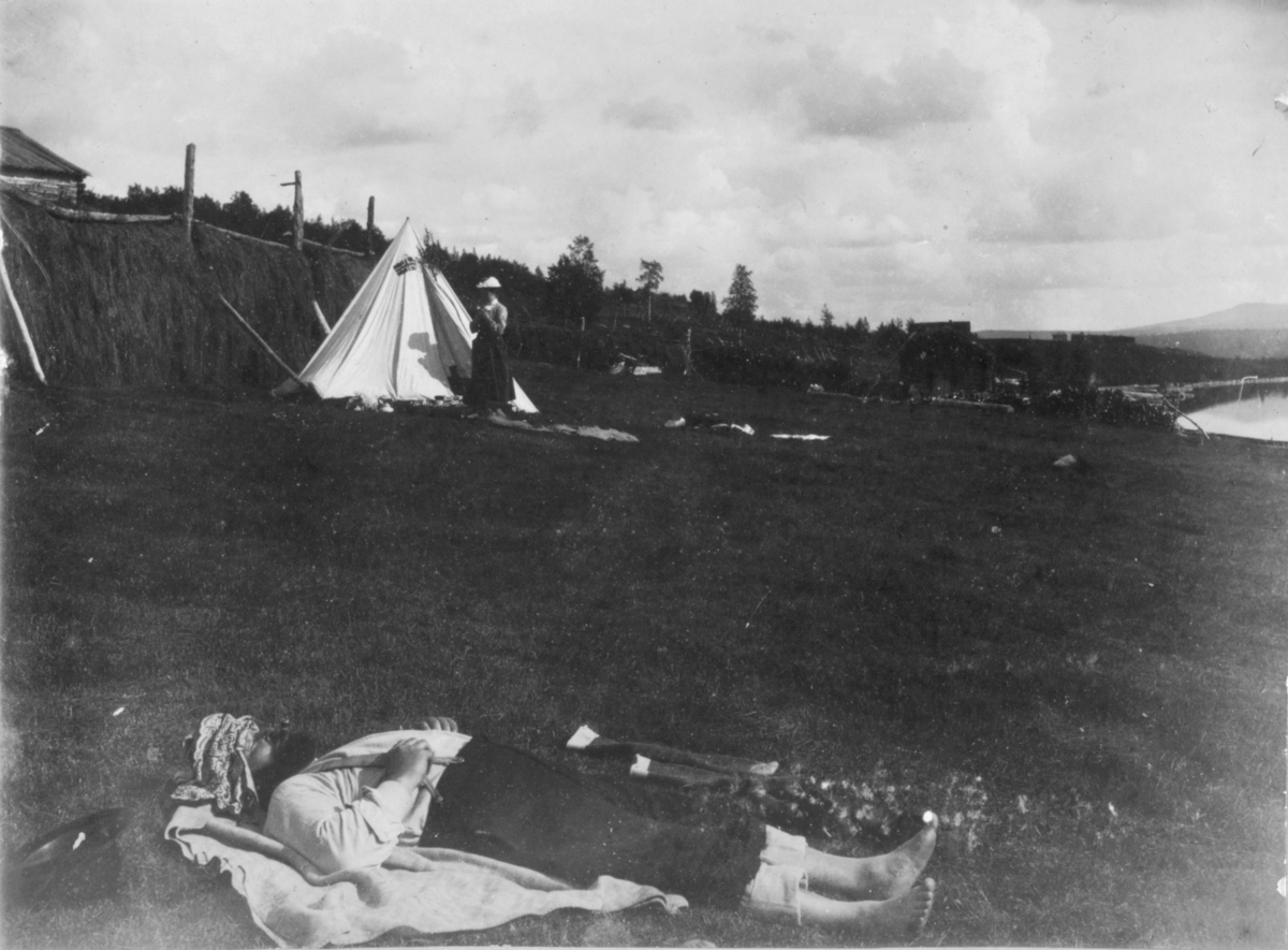 En mann ligger på bakken og hviler. Han har et tørkle over ansiktet. I bakgrunnen et telt med en dame i teltåpningen. Hun har side skjørt og en lys hatt på hodet.