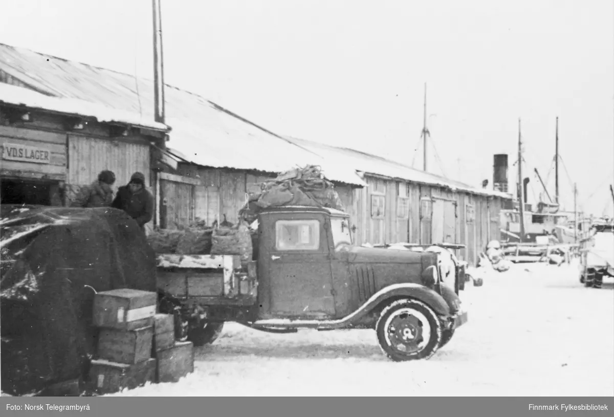 'Vadsø har likevel en av de beste havner i Finnmark. Her lossesforsyninger og bygningsmaterialer til hele distriktet.' På den enestebrukbare kaia er det satt opp lagerbrakker. Her er to menn i ferd medå laste på en lastebil, en Chevrolet 1934-35 modell. En av brakkene har et skilt merket 'vdslager'.