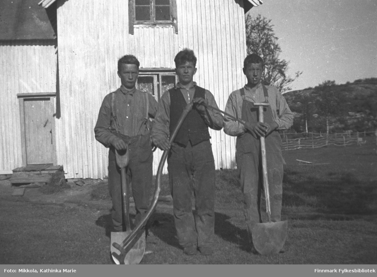 Tre unge menn med spader og ljå, fotografert foran et lyst hus. Vi vet ikke hvor bildet er tatt, men det er antakelig i Sør-Varanger