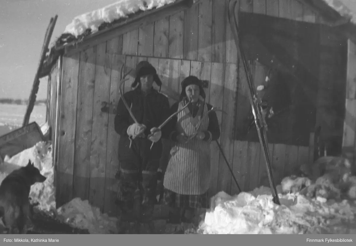 Ved et uthus i Neiden. Til venstre Arvid Mikkola, til høyre Helga Hallonen, begge holder reingevir fram mot fotografen. I snøen står et par ski, til høyre i bildet en hund