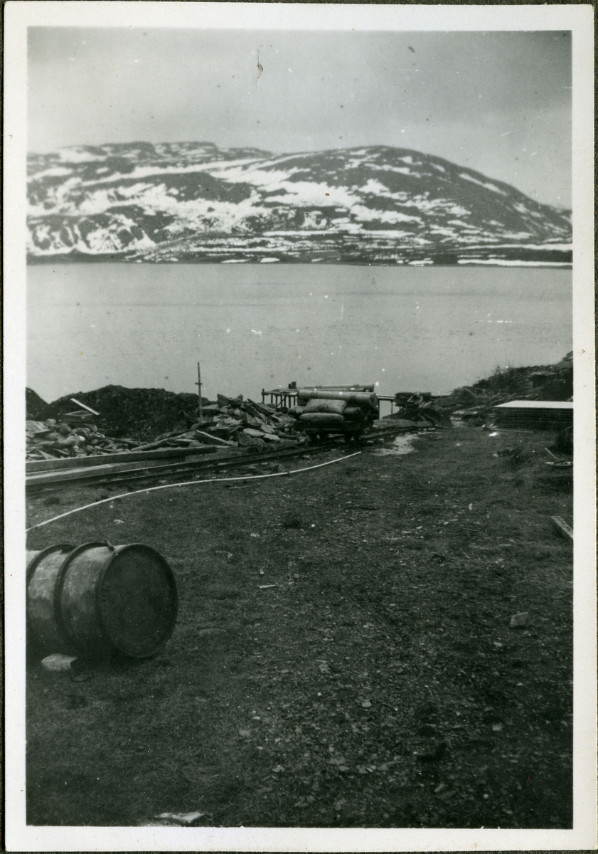 Fotografi av jernbaneskinner med vogn. Vognen er lastet med byggemateriale og hører til skiferbruddet i Friarfjord. I forgrunnen kan man se en stor tønne. I bakgrunnen ser man havet og fjell.