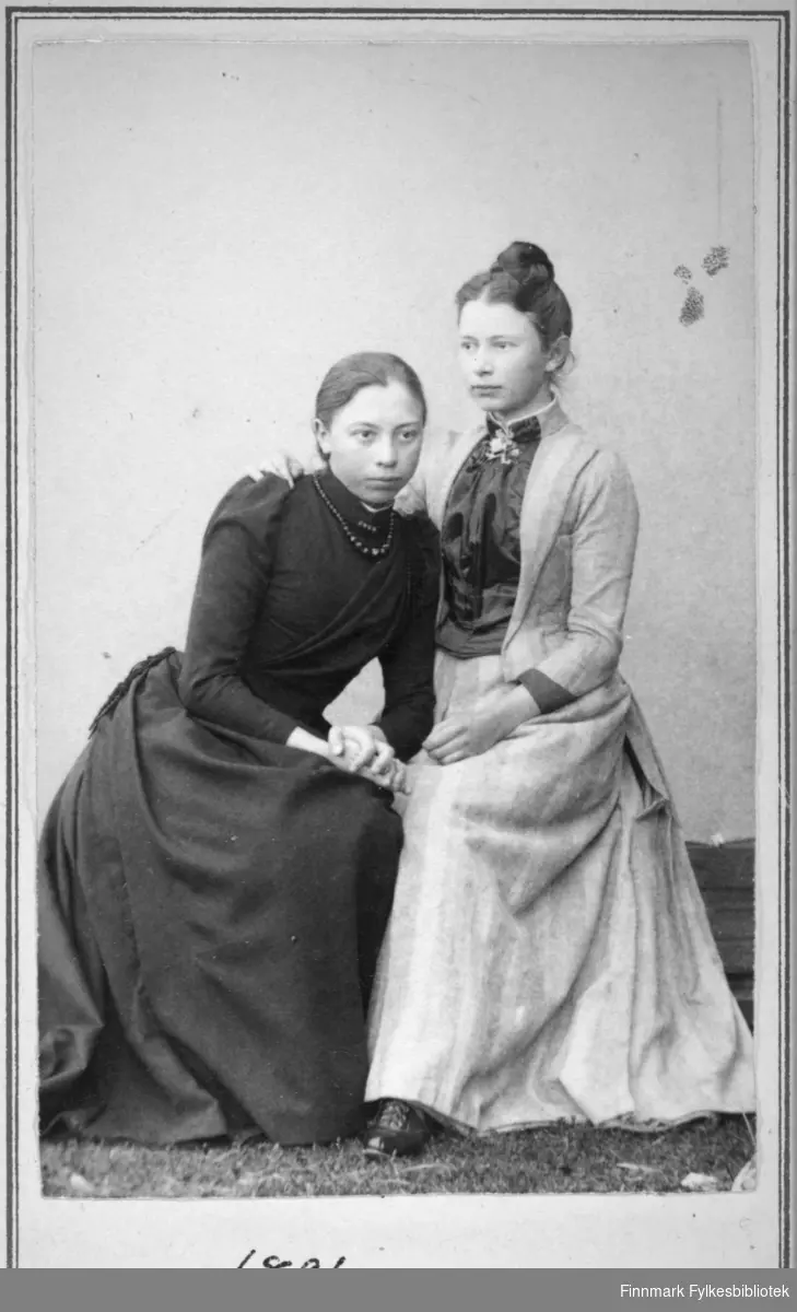 Portrett av to unge damer. Damen til venstre på bildet har en sid, mørk kjole på seg. Damen til høyre på bildet har en lysere kjole med et stort smykke/brosje i halsen. Portrettet er tatt hos fotograf Kalland i Hammerfest.
