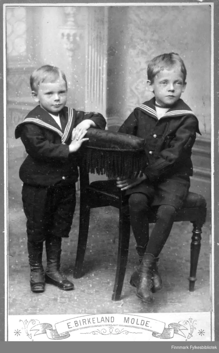 Portrett av to gutter. Den ene sitter i en stol, den andre lener seg mot stolen. Begge har mørke kortbukser, mørke strømper og sko. De har også hver sin like matrosjakke. Tapetet bak dem har interiørmønster.