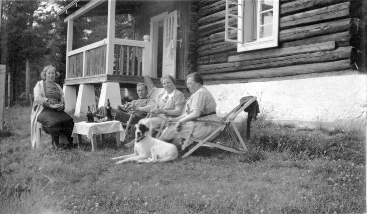 Tre damer, en mann og en hund koser seg i hagen utenfor et hus eller hytte.