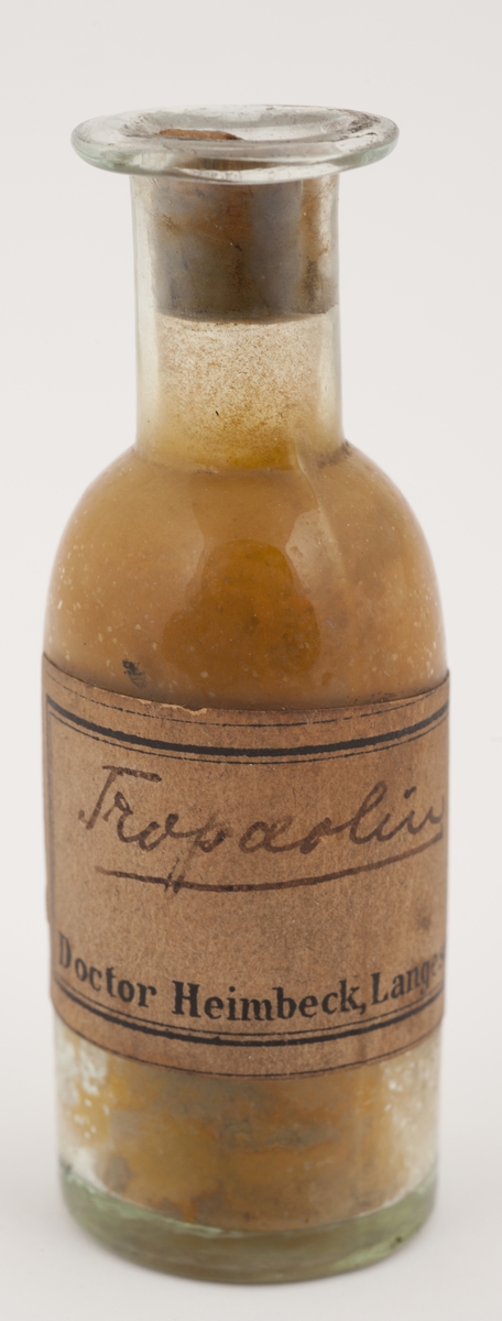 Rund, klar flaske med propp av kork. Innstøpt i glasset er mål på 5 - 10 - 15. Inneholder  lysebrunt pulver.