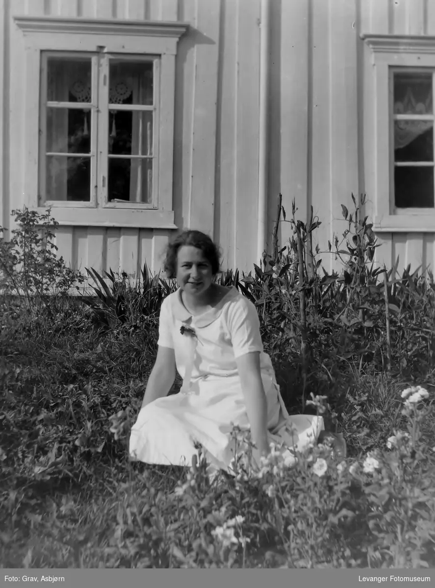 Kvinne i lys kjole sitter foran hus.