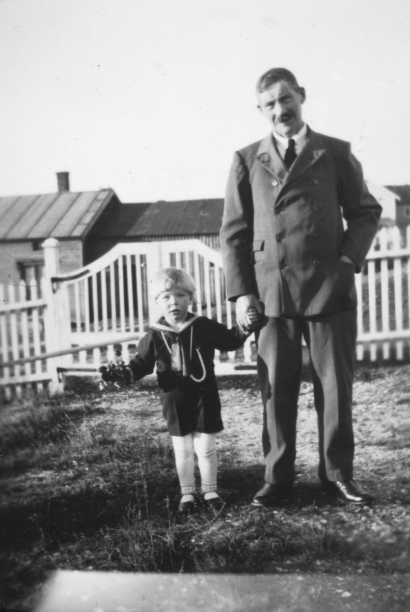 Tor Hauge, og bestefaren Alfred Amundsen står hånd i hånd, i hagen i Nyborgveien. Bildet er muligens tatt 17. mai. Alfred er kledt i dress, og Tor har på seg en matrosdress. Bak porten og gjerdet ses Vilmigården