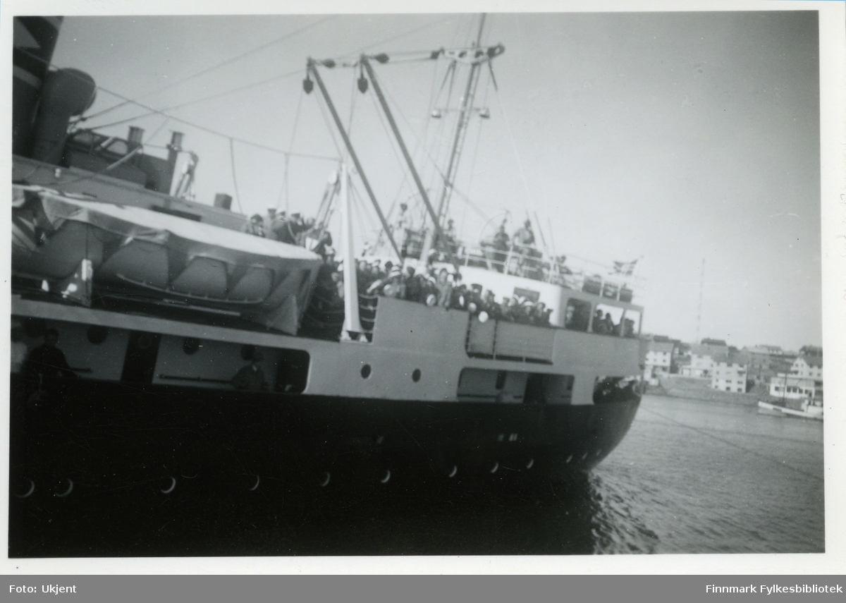 Bildet trolig tatt av et medlem i Vadsø damekor. Medlemmer av Vadsø damekor er muligens ombord på skipet. På båten henger det en livbåt. Det er mange mennesker på dekk. Til høyre kan man se bebyggelse. Skipet er hurtigruten MS Barøy,som var i drift fra 1953 til 1964 og var eid av Ofotens Dampskipsselskap, Narvik.