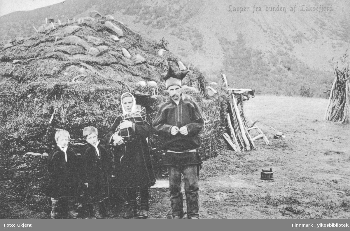 Påskrift på postkortet: 'Lapper fra bunden i Laksefjord'. På bildet står det som trolig er en familie: to unge sønner, mor med et spedbarn i hendene og far. De er kledd i kofter, skaller og samiske klær. I bakgrunnen kan man se en gamme og på bakken kan man se et trespann. Kvinnen har et sjal på hodet, mannen en lue. Postkortet er datert julaften 1921.