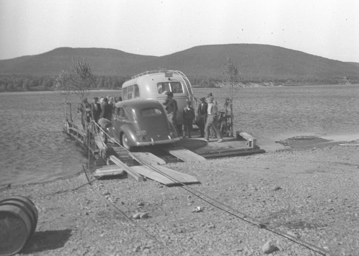 Flere personer, en personbil og en minibuss ombord i kabelferga som gikk over Tana-elva ved Seida. Bussens registreningsnummer er muligens Y-2142, som var en Volvo personbuss, 1939-modell, som tilhørte A/S Polarbil i Vadsø. Polarbil hadde ruter i Øst-Finnmark inkl. Tana.
