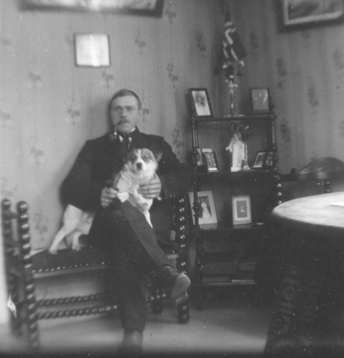 Reidar Håheim poserer for et bilde med hund i fanget. Reidar er kledd i dress og slips og har en pipe i munnen. Bak han kan man se en hylle med fotografier og et norskeflagg.