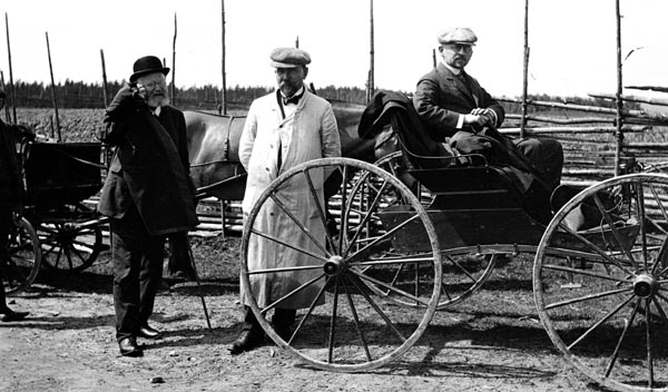 Direksjonen (styret) i Christiania Tømmerdirektion på befaring.  Reisa foregikk med hest.  På dette fotografiet poserer tre av styremedlemmene, to stående og den tredje sittende på en karjol. Mannen til venstre er Bernhard Magnus Kopperud. Han som sitter i karjolen er muligens Otto Nilsen.
