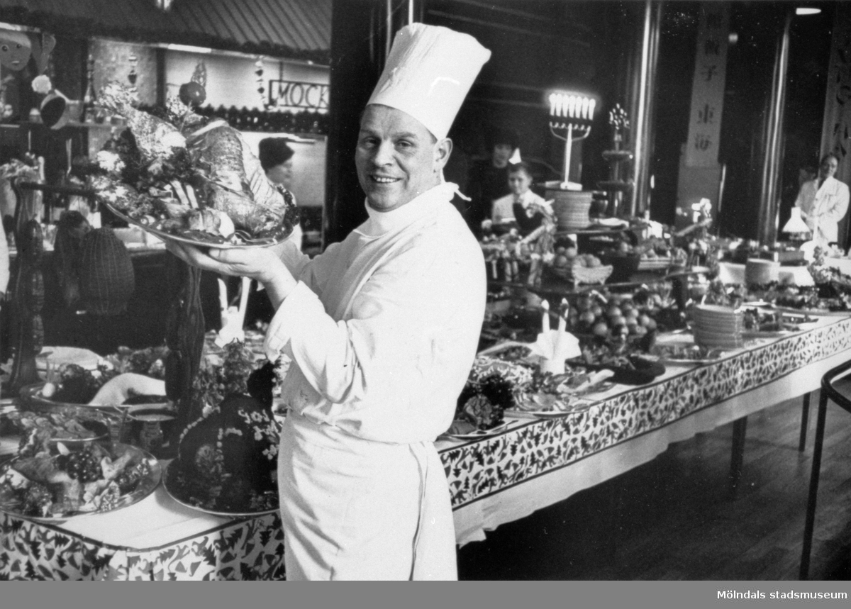 Gunnebo slotts egen kock Ruben Bohm (anställd 1938-1943). Han står med ett fat i handen bredvid ett långbord fyllt med mat. I bakgrunden ses serveringspersonal, 1940-tal.