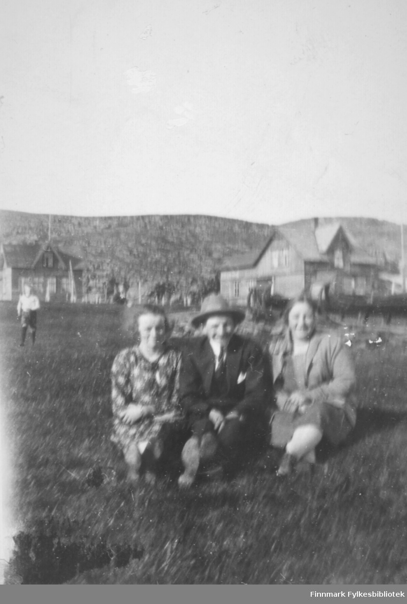 Tre personer sitter i gresset en sommerdag i Kiberg (hvis ikke i Hamningberg?) før krigen. Fra venstre: Aline Strige, muligens Aksel Georg Johansen i midten fra Hamningberg (eller Havningberg som det hette den tiden) og Gudrun Bauna til høyre. 