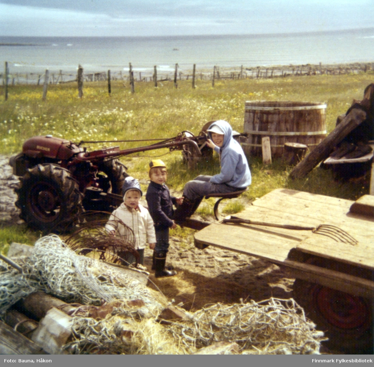 Tre barn ute på et jorde med mye redskaper rundt dem, bl.a Arnulf Baunas traktor. Til venstre står Elin Bauna, de to guttene er ukjent.
