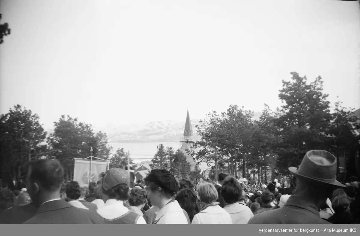 17. mai feiring på Elvebakken i alta. Kirkespiret på Elvebakken kirke og Altafjorden kan ses over folkemengden.
Bildet er tatt på våren 1962.