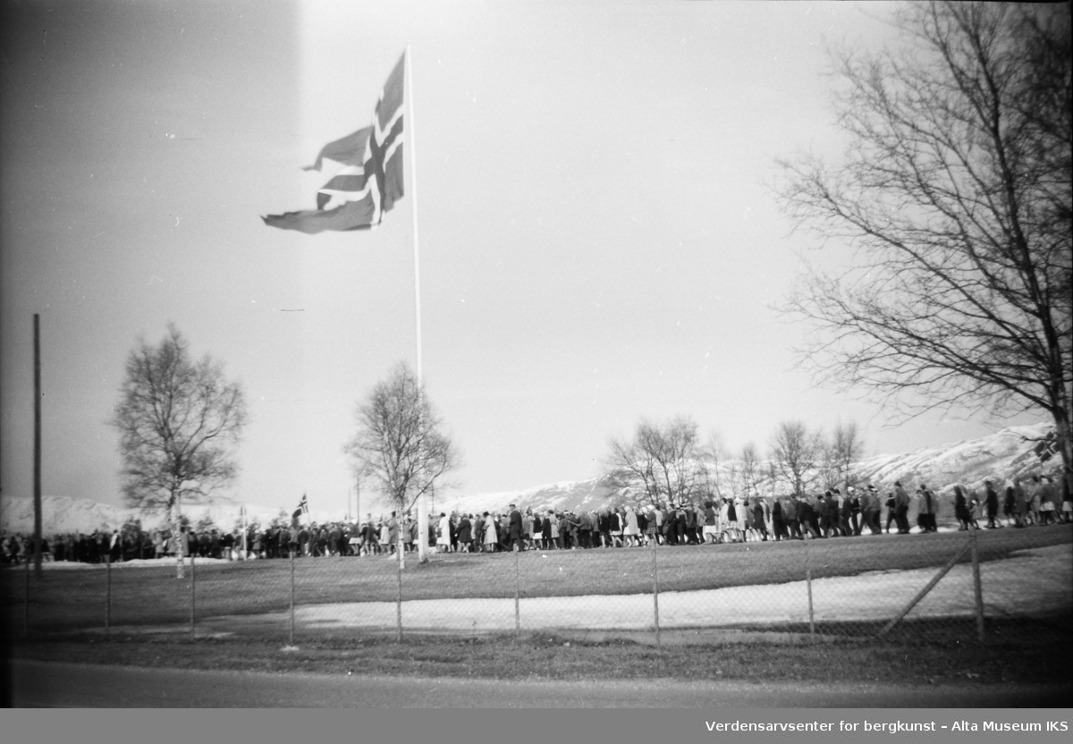 Et tog av mennesker går forbi en flaggstang på 17. mai. Et stort norsk flagg vaier i vinden fra toppen av flaggstangen.
Bildet er tatt på våren 1962.
