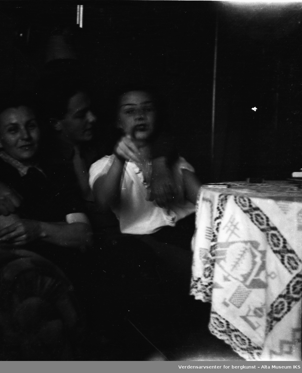 En ung mann og to unge kvinner sitter i en sofa. Mannen har hånden rundt en av kvinnene, som peker mot kameraet.
