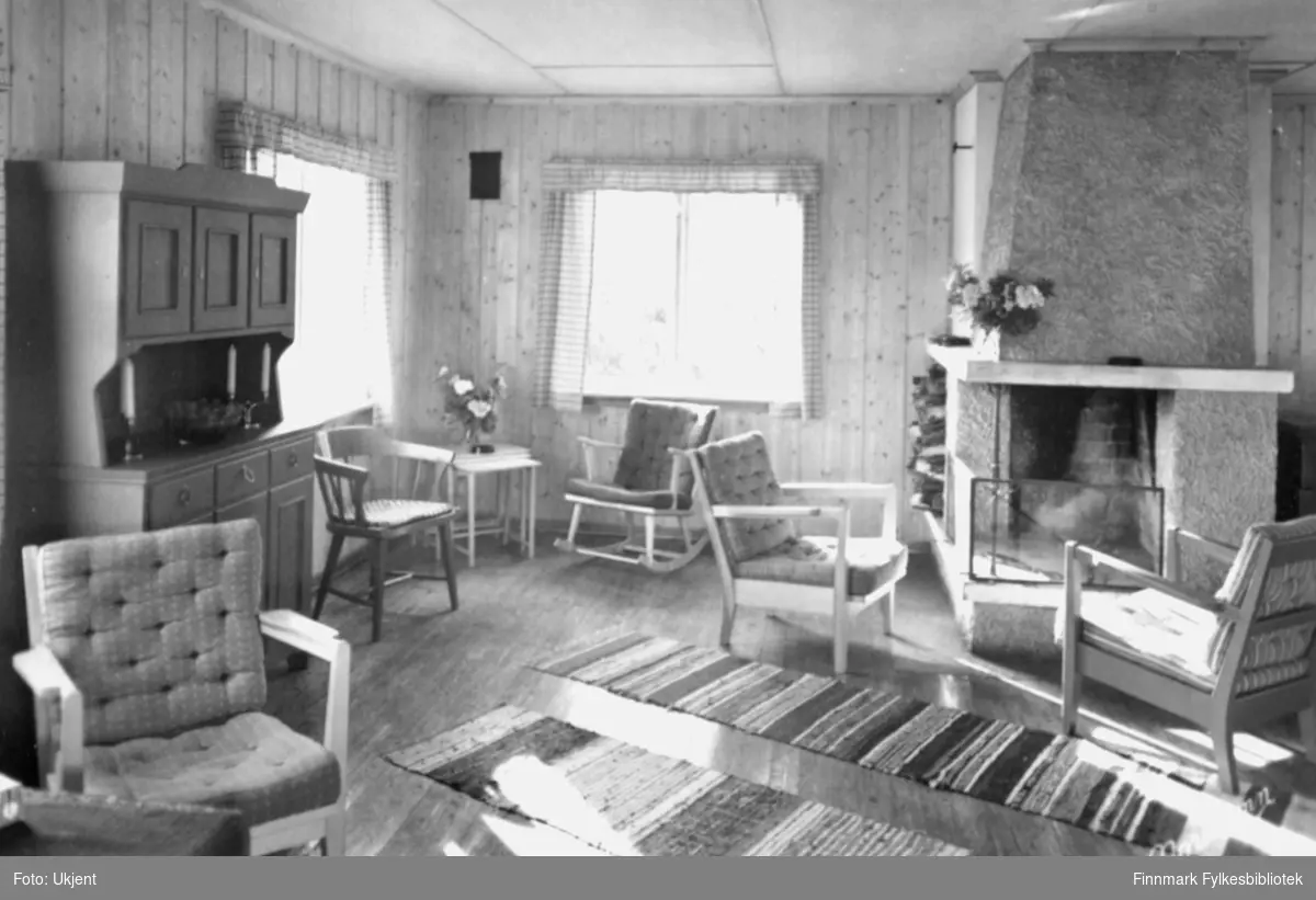 Et bilde tatt inni Ifjord gjestgiveri. På bildet kan man se stoler, matter, vindu, gardiner, ildsted, skap, talgelys og blomster.  Bildet er trolig tatt i løpet av siste halvdel av 1900-tallet.