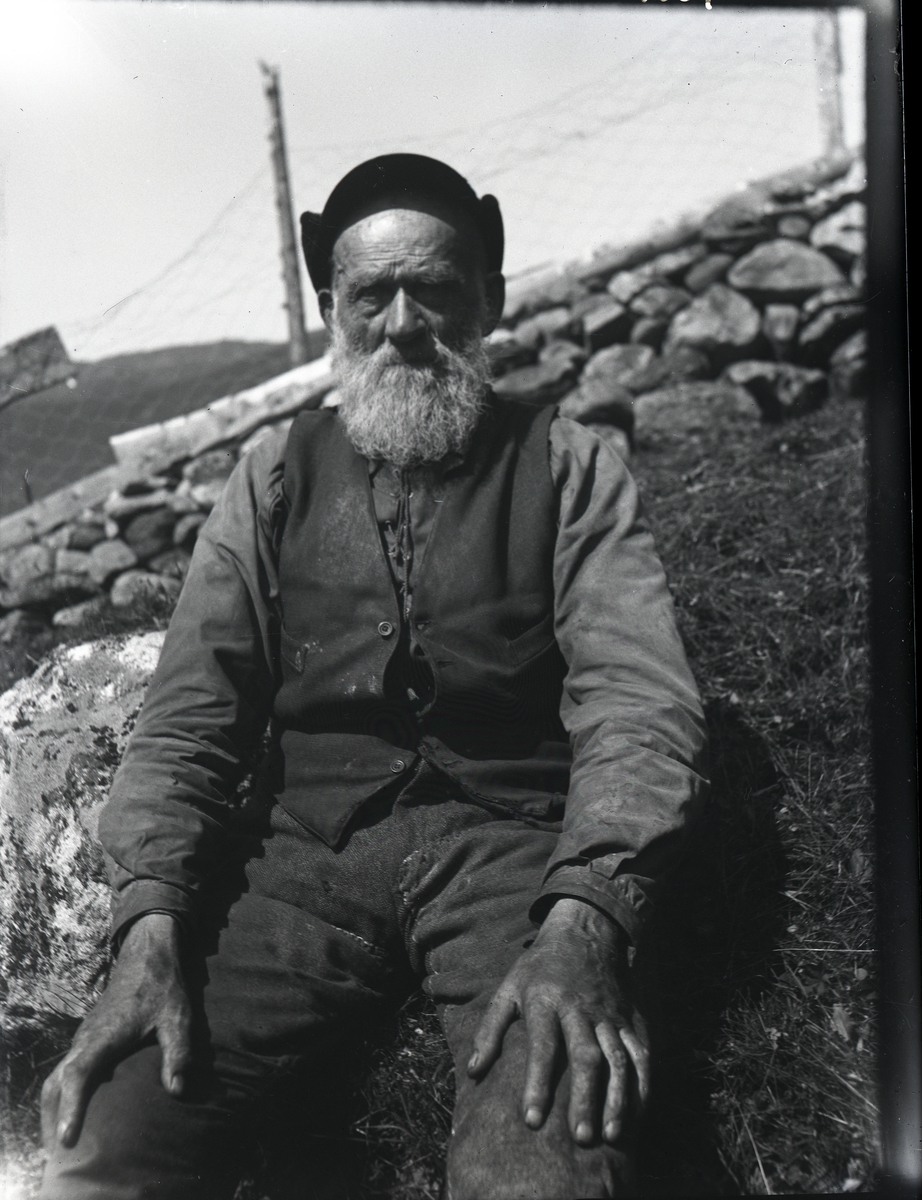 Mann som sitter på grasbakken foran en steinmur. Han har hvitt skjegg og er iført arbeidsklær; skjorte med snøring, vest og en lue/hatt på hodet.