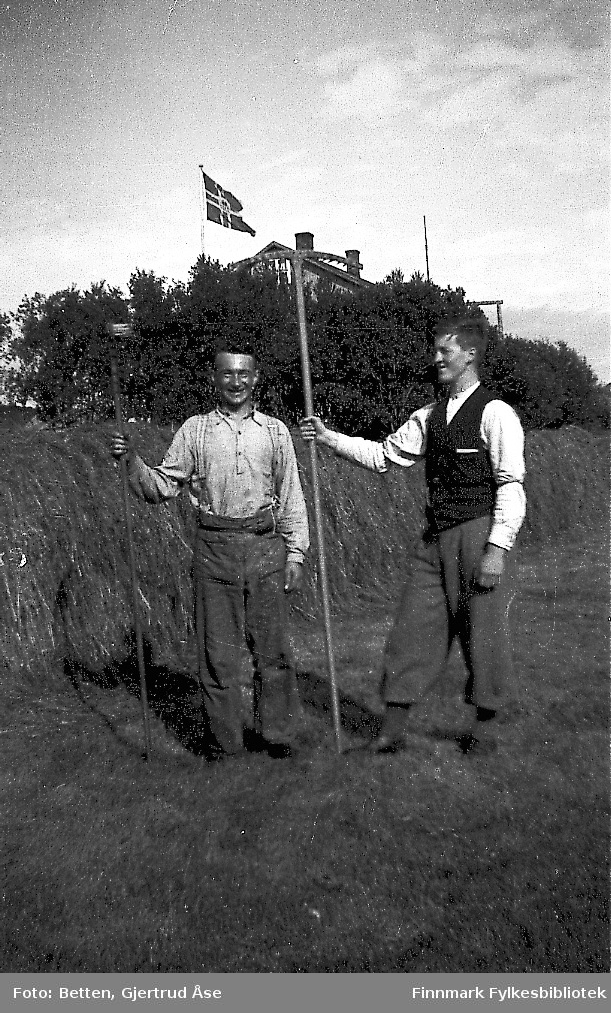 To menn, Isak og Einar, står på slotta med en ljo og en rake på hand. Høn er berget og hengt til tørk. Bakerst på bildet står et hus bak trår. Den norske flaggen er heist opp.