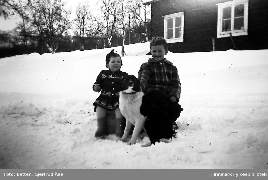 To barn, en jente og en gutt, sitter på en trekasse på vegkanten ved et bolighus. Mellom dem sitter en sort/hvit hund. Den lille piken på ca 2 år heter Svanhild Johansen og gutten på 6 år Bjørn Ottar Betten. Landskapet er dekt av snø. Bakerst står huset.