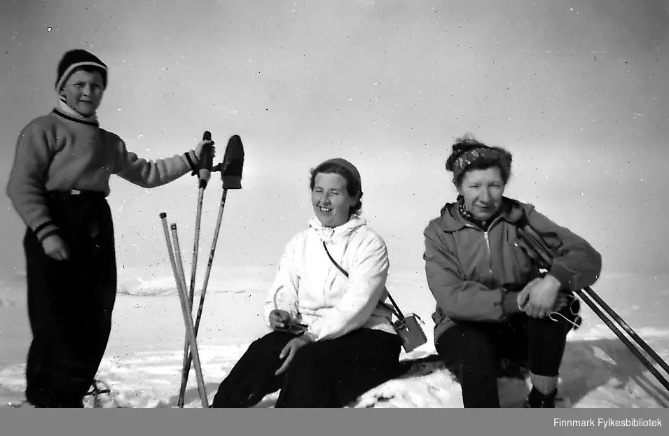 En gutt og to kvinner på skitur i Seida en vakker og solig påskedag. Gutten er kledd i en gensere og en lue. Fra venstre: Rudolf Betten, hannes tante Gjerdrud Betten og Åsta Mickelsen sittende på snøen ved gutten.  Kvinnene har på seg skibukser og anorakk. Et par skistaver står i snøen