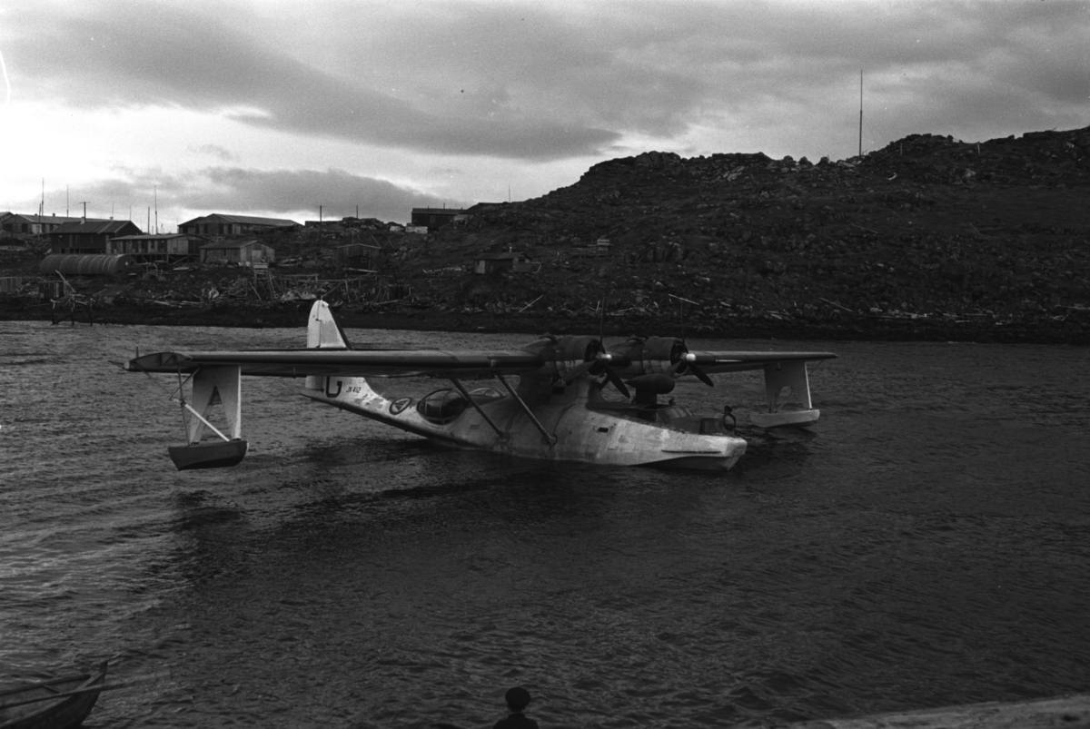 En flybåt av merket Catalina Mk IVB har landet i Honningsvåg like etter andre verdenskrig. På halepartiet kan en lese JX 412 og en stor G,( KK-G). Flyet fikk ny bokstavkode i løpet av 1946 til K-AK. Ifølge fotografen er bildet tatt i 1946 med gammel bokstavkode G som bildet viser. er et fly som starter og lander på sjøen. 

En flybåt skiller seg fra et sjøfly ved at selve skroget flyter på vannet, skroget er dermed formet som på en båt. Eksempler på flybåter er for eksempel Short Sandringham og Dornier Do X. Flybåtene var svært populære før nettet av flyplasser var skikkelig utbygd, og egnet seg bedre for langdistanseflygninger enn samtidige landbaserte passasjerfly.

Arkitekt Ola Hanche-Olsen arbeidet ved Brente Steders Reguleringskontor i 1946. Hovedadministrasjon for gjenreisning av Nord-Troms og Finnmark ble lagt til Harstad og fikk navnet Finnmark kontoret. Landsdelen Nord-Troms og Finnmark blev oppdelt i syv distrikt med hver sin administrasjon. Honningsvåg, distrikt IV, skulle betjene Nordkapp, Lebesby, Porsanger og Karasjok kommune.

Ola Hanche-Olsen har tatt bildene. Han var født 13. mars 1920 i Borre, død 11. februar 1998 i Gjettum. Han var både arkitekt og barnebokforfatter. Han hadde artium fra 1939, arkitekteksamen fra NTH 1946 og arbeidet deretter ved Finnmarkskontoret 1946–48 før han etablerte egen arkitektpraksis. Han debuterte som barnebokforfatter i 1974 med lettlest-boka "Knut og sjørøverne", og skrev i alt 12 bøker. Han var XU-agent 1944-45, og var også en aktiv fjellklatrer og friluftsmann. Ola var gift med Solveig Hanche-Olsen (f. Falkenberg); de fikk 3 barn, blant dem matematikeren Harald Hanche-Olsen.

