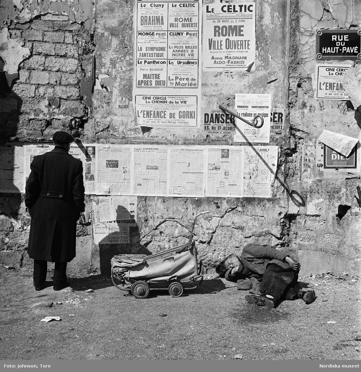 Paris, Rue du Haut-Pavé. Två män, en stående läsandes en väggtidning, den andra sovande. En dockvagn i mitten.