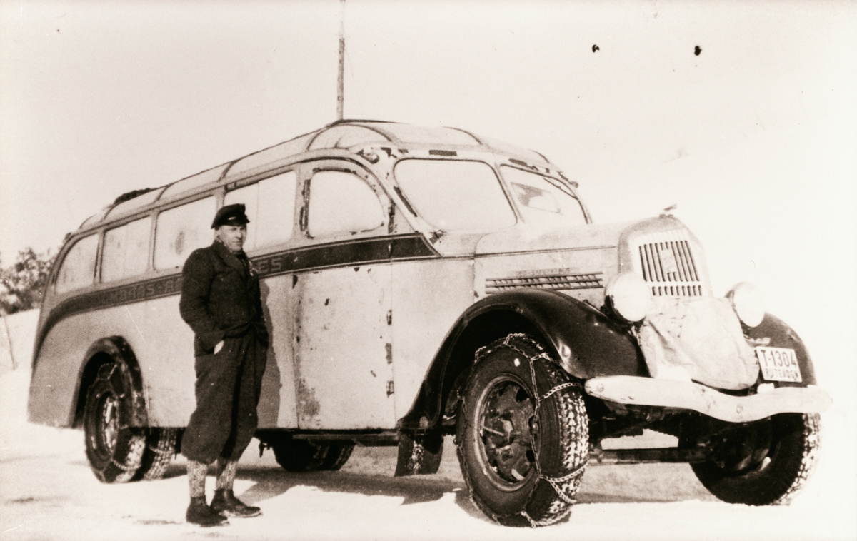 En mann som er sjåfør står foran en rutebil med kjettinger på dekka. Veien er snødekt. Rutebilen var den første som ble brukt på ruta mellom Åndalsnes og Dombås ved starten i 1941.