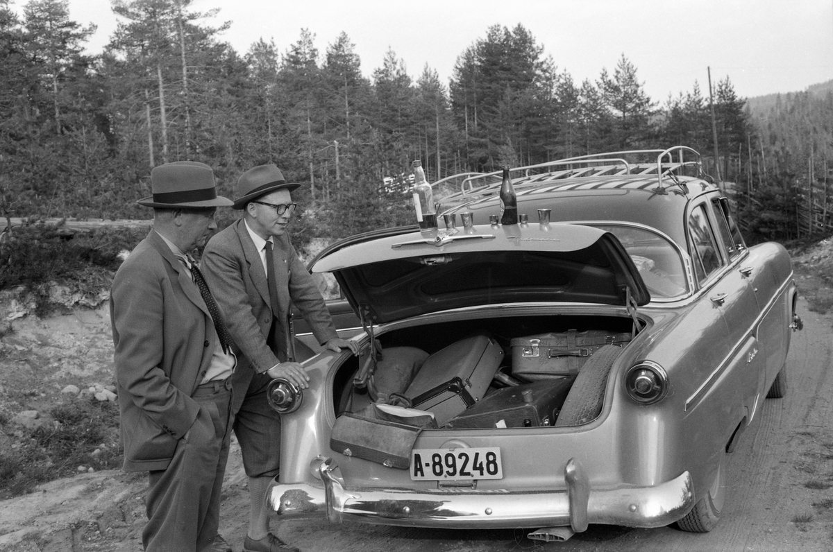 Fra Glomma fellesfløtingsforenings direksjonsbefaring 26. mai 1960. Fotografiet er tatt ved en sideveg i Gudbrandsdalen. Ekskursjonsdeltakerne hadde åpnet bagasjelokket på bilen og satt to flasker og fem drammeglass der. I sjølve bagasjerommet lå ekskursjonsdeltakernes kofferter. To av dem sto ved bilens venstre bakskjerm. Kjøretøyet var en amerikansk 1954 Ford Customline (evt. Crestline) med automatgir.
