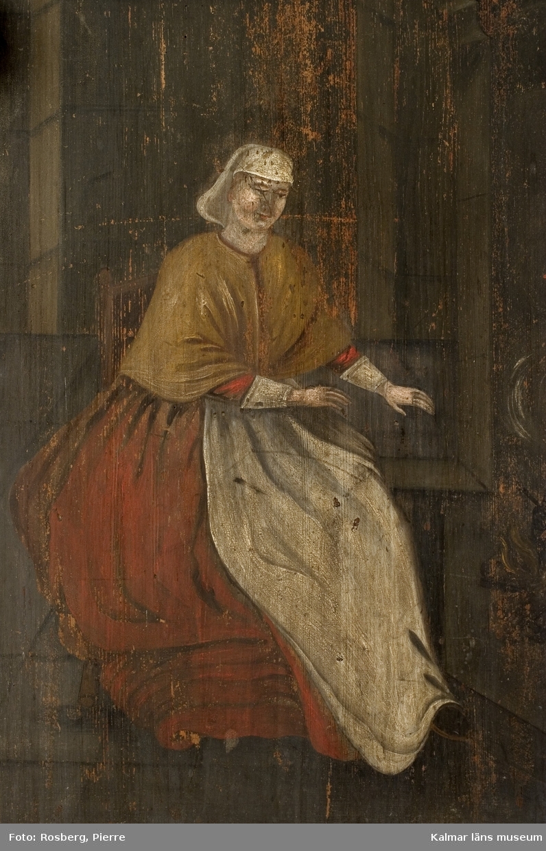 Porträtt av kvinna, helfigur, interiörmotiv, kvinnan sitter framför spisen med eld och kokkärl.