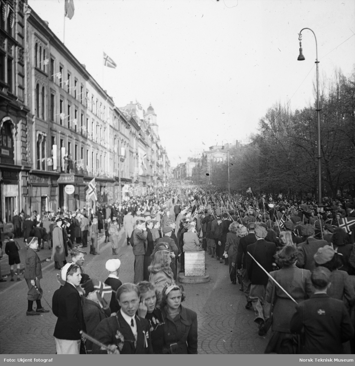 To jenter og en gutt pyntet med norske flagg og 17.mai-sløyfer foran russetoget som passerer stor folkemengde på Karl Johan mot Egertorget, Oslo, mai 1945,