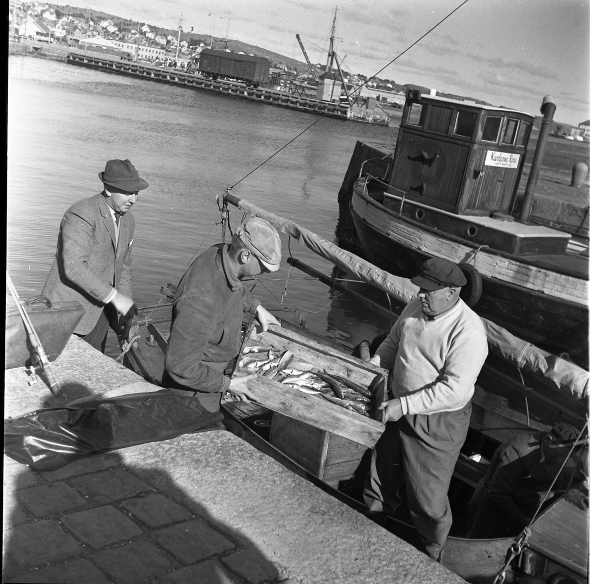 En inombordare ligger vid kaj. En låda med fisk lyfts upp ur båten av två män. På kajen ligger ett kastspö över en portfölj.