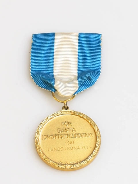 Medalj med texten :"LANDSKRONA IDROTTSFÖRENINGARS SAMORGANISATION" som omringar Landskronas stadsvapen på ena sidan och texten: "FÖR BÄSTA IDROTTSPRESTATION 1961 LANDSKRONA G.I.F." på andra sidan. Stämplar : "RW", (oläsbar), "S", kattfot, "M9". M9 betyder 1960. Hänger i blått och vitt band, fästes på kläder med säkerhetsnål.