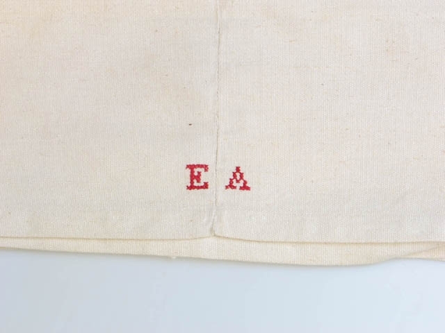 Grovt linnelakan, ihopsytt av två delar. Längst ner monogrammet "EA" broderat i rött med korsstygn. Elin Andersson (1874-1929) var en av systrarna Andersson på Asmundtorp 7 och 9 (Utställningen Systrarna från Asmundtorp). Fyra av de sex systrarna gifte sig inte och deras hemgifter, som bestod av nio kistor med egentillverkade textilier, blev inte använda.
