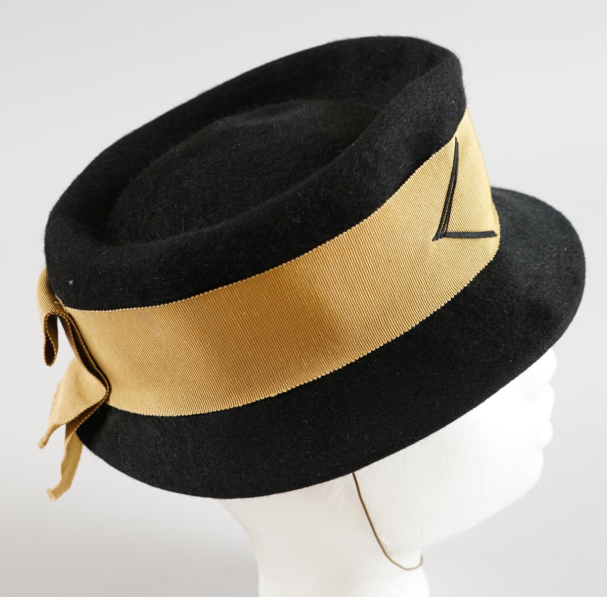 Svart hatt i ullfilt med gyllent pynte band og tynt strikk til feste.