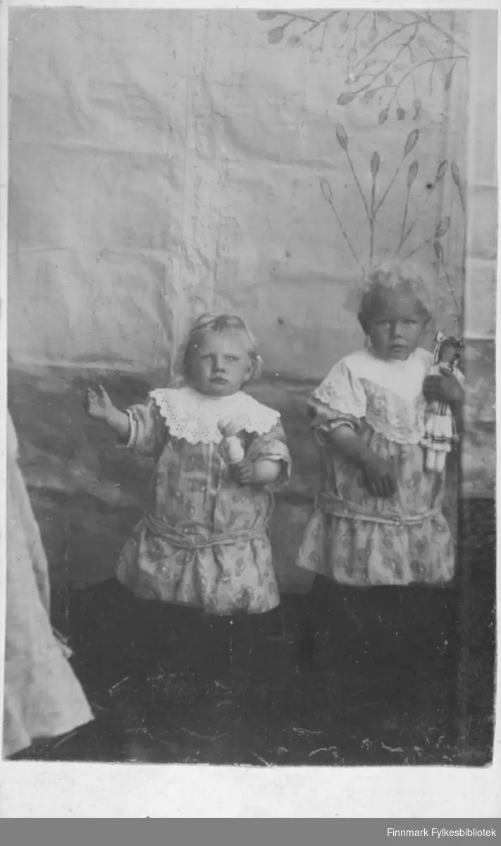Søstrene Else Sofie (1909-1913) og Thora Johanna (1910-1972) Pedersen, Sørvær, Helligvær, Nordland. Jentene er kledd i fine kjoler med krage og begge har dukke i hånden, fotografert i fotoatelier. 