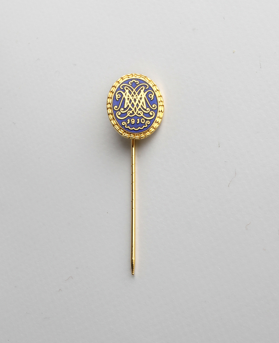 Jakkenål, gullfarget, oval form. Ei lang nål er festet til baksiden.

Motivet er Den norske Amerikalinjens logo i gull på mørk blå bakgrunn, med årstallet 1910.
