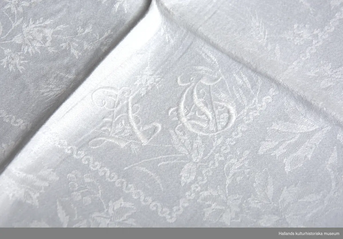 En vit linneduk med tillhörande servetter. Föremålen har en dekor av sirliga mönster kombinerat med en flora av örter och lövverk. Samtliga föremål är broderade med signaturen "HG" (Hugo Gerlach). Det finns ytterliggare en signatur på duken - se bild 2.
