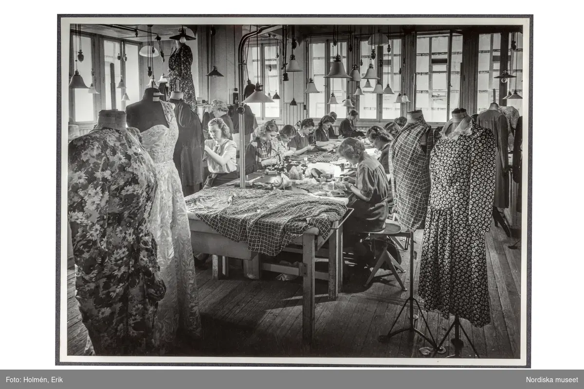 Interiör från Nordiska Kompaniet. Sömmerskor arbetar vid ett bord i NK:s Franskas damskrädderis ateljé. I förgrunden syns kläder på provdockor.
