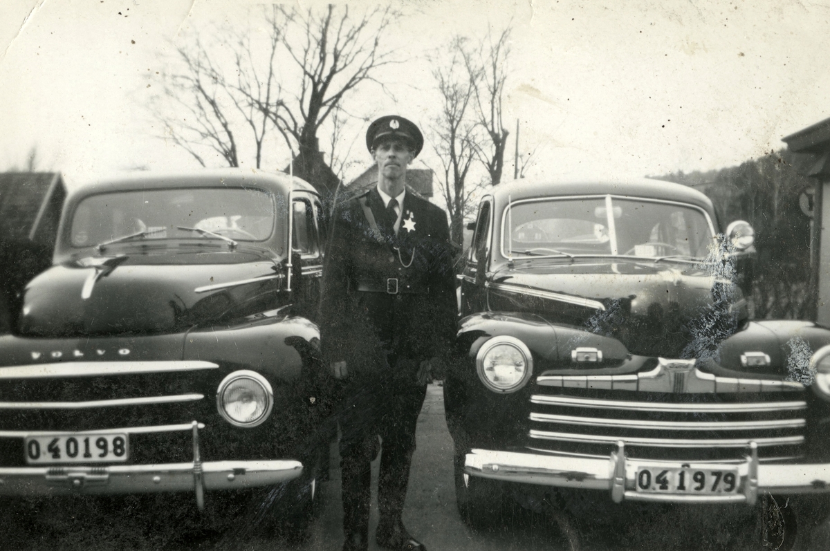 Fritiof (elev boende på Stretereds skolhem) iförd polisuniform, står mellan två taxibilar vid Kållered station, 1940-tal. Han fick "hjälpa" polisen ibland. Taxistationen skymtas till vänster. Relaterat motiv: A2301.